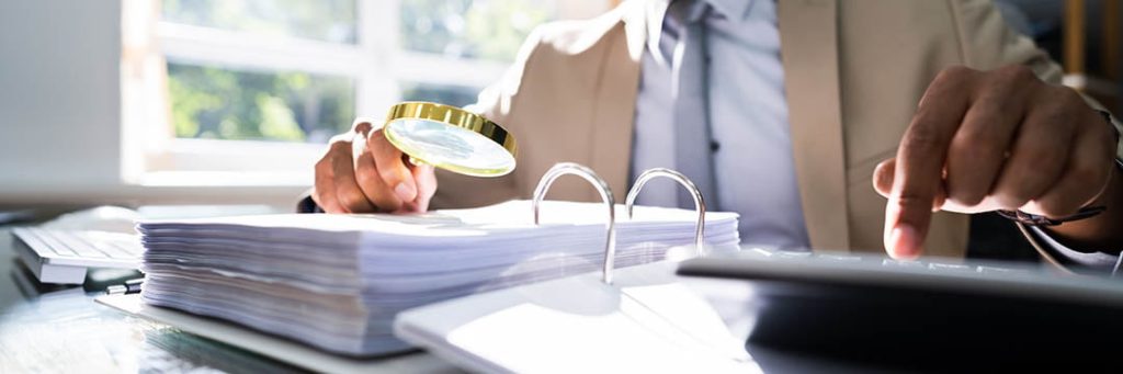 Man in a suit examines paperwork with a magnifying glass while entering numbers into a calculator